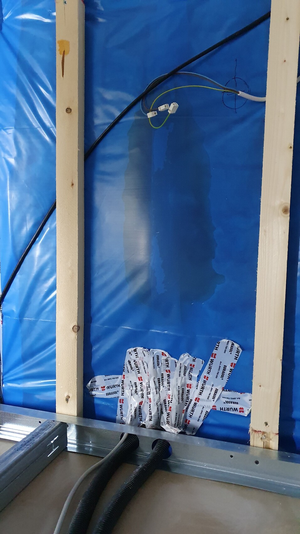 Bauwand mit blauer Folie, Holzlatten, sichtbare Kabel und flexible Rohre am Boden.