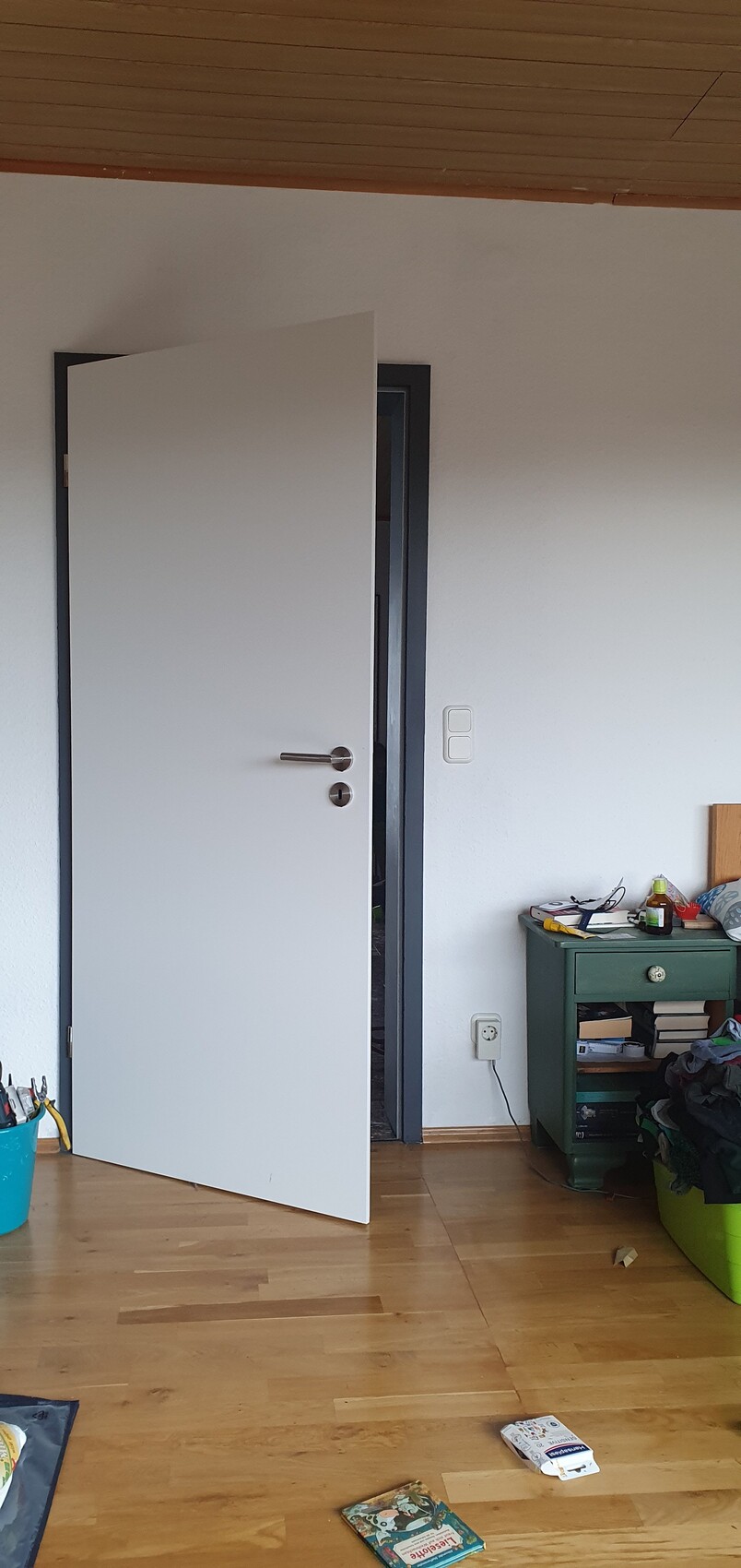 Offene Zimmertür in hellem Innenraum mit Holzfußboden, grünem Sideboard und chaotischen Gegenständen.