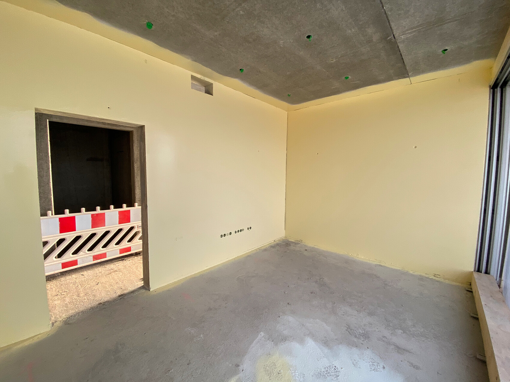 Unfertiger Innenraum mit gelben Wänden, rohem Betonboden und Tür zur Baustelle mit Absperrband.