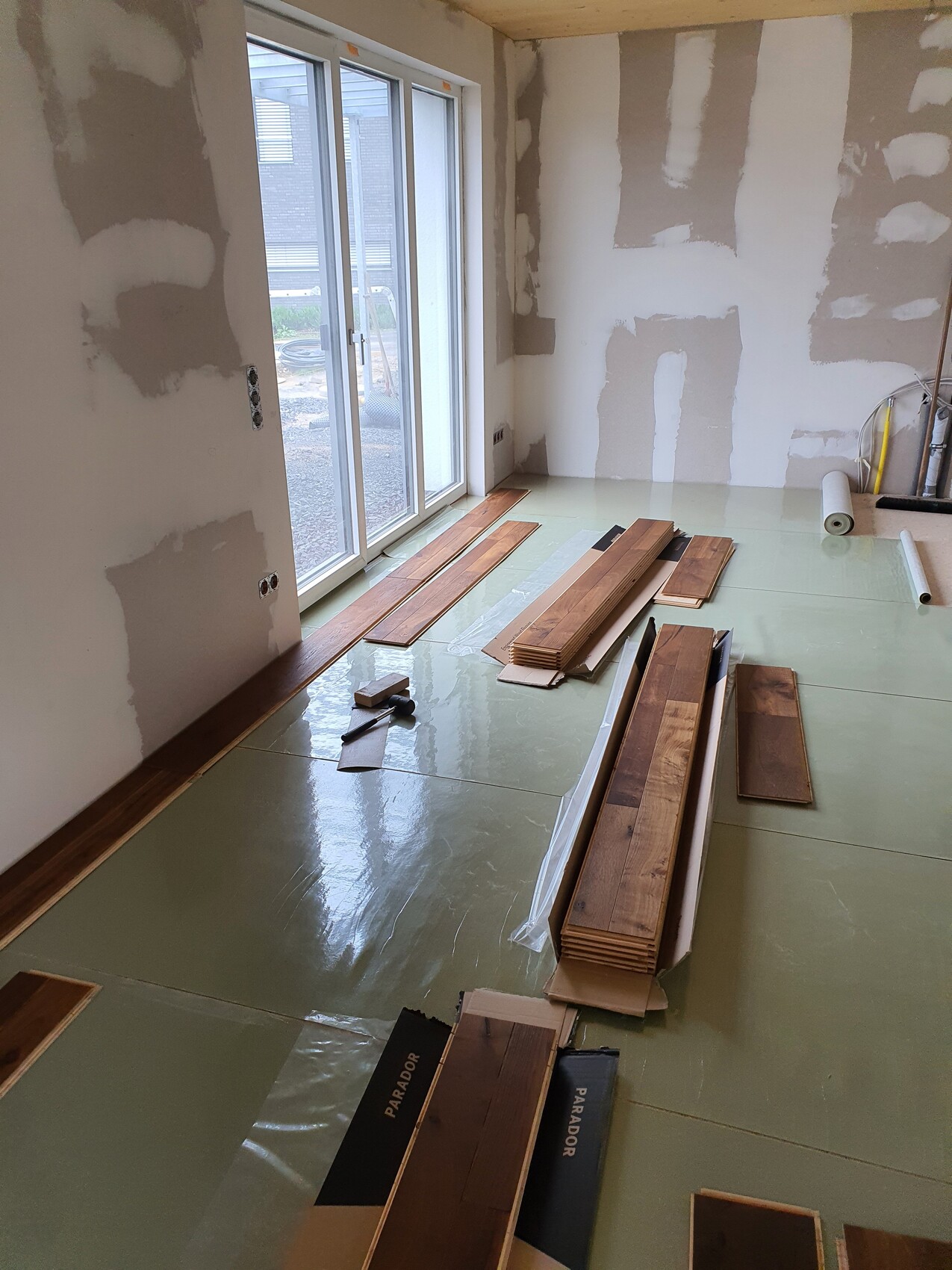 Renovierungsraum: Glas-Schiebetür, verstreute Holzplanken, Bauholzstapel auf grünem Unterboden.