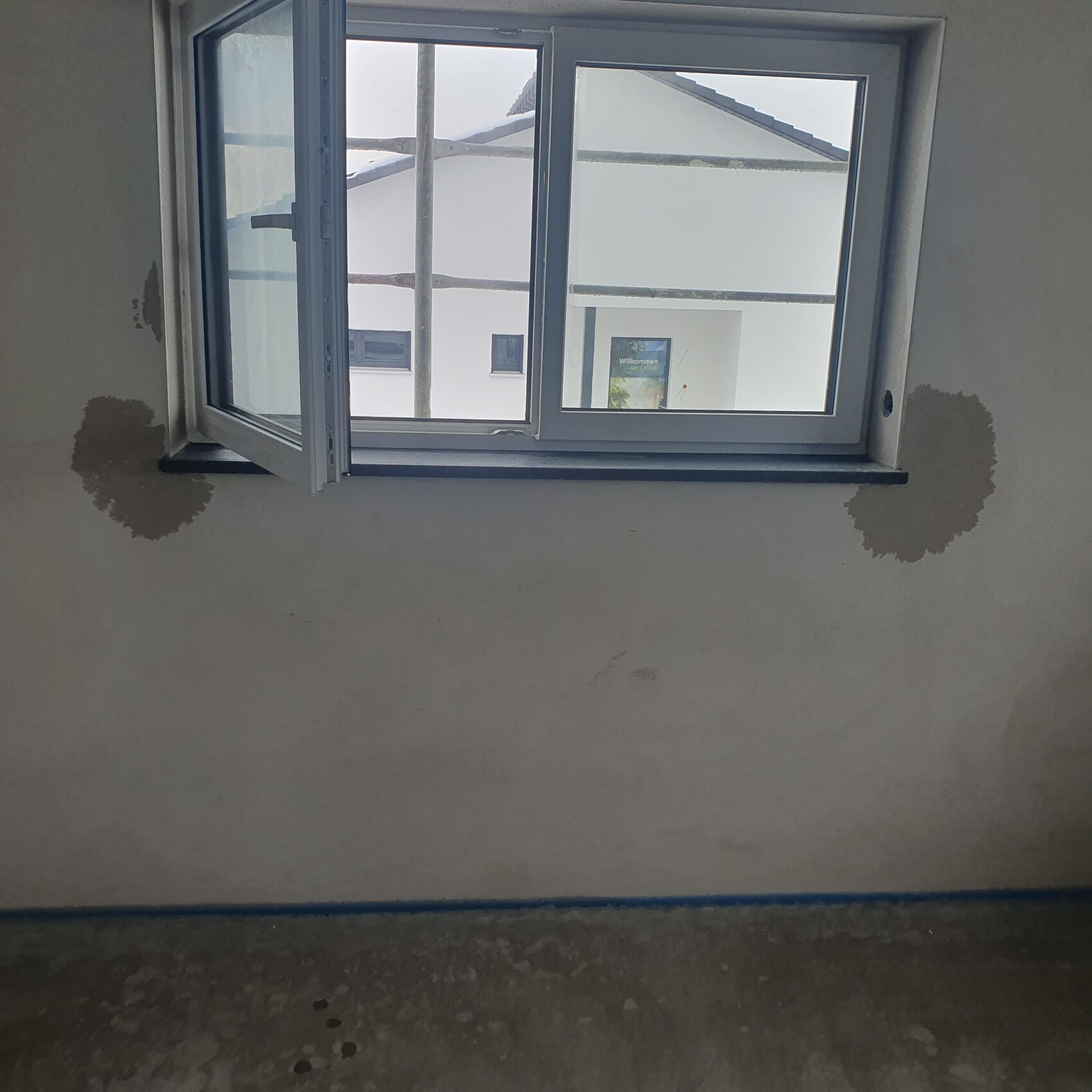 Offenes Fenster in unfertiger Innenwand; Putzstellen, Betonboden, Blick auf weiße Fassade.