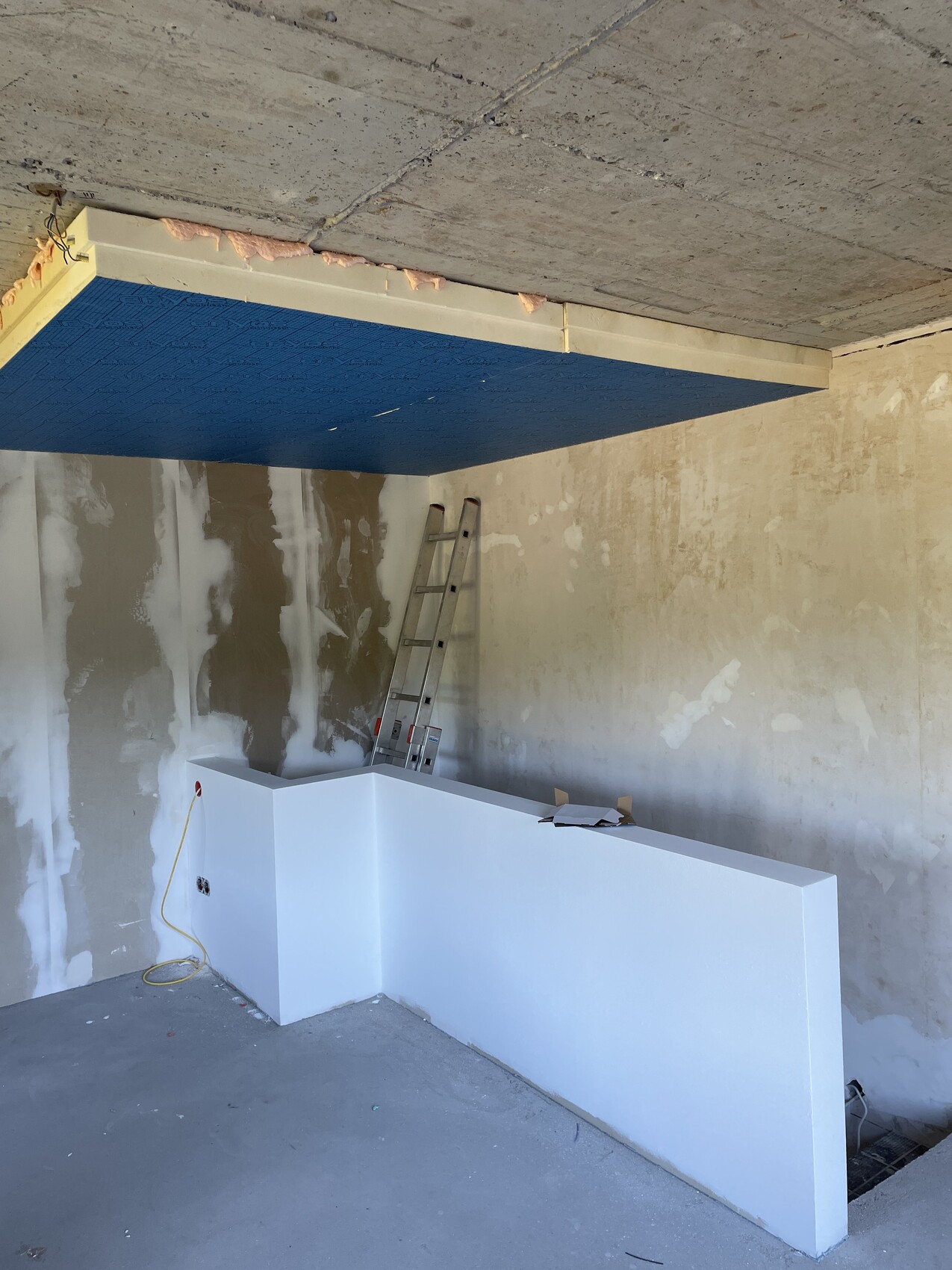 Baustelle: weiße Bar/Theke vor unfertigen Wänden, Leiter, blaue Deckenplatte, Kabel am Boden.