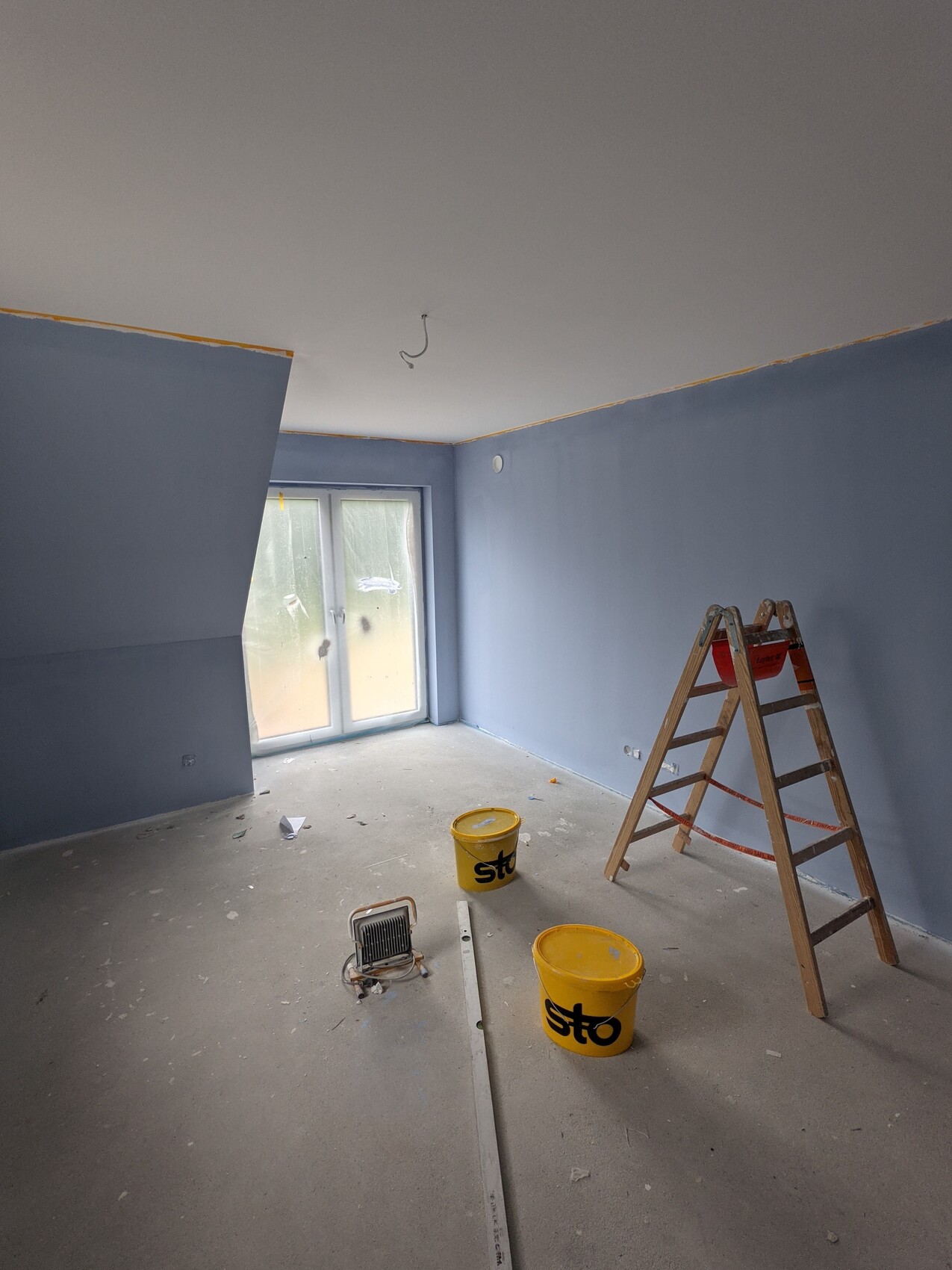 Renovierungszimmer mit blauen Wänden, Leiter, gelben Sto-Eimern und Fensterfront.
