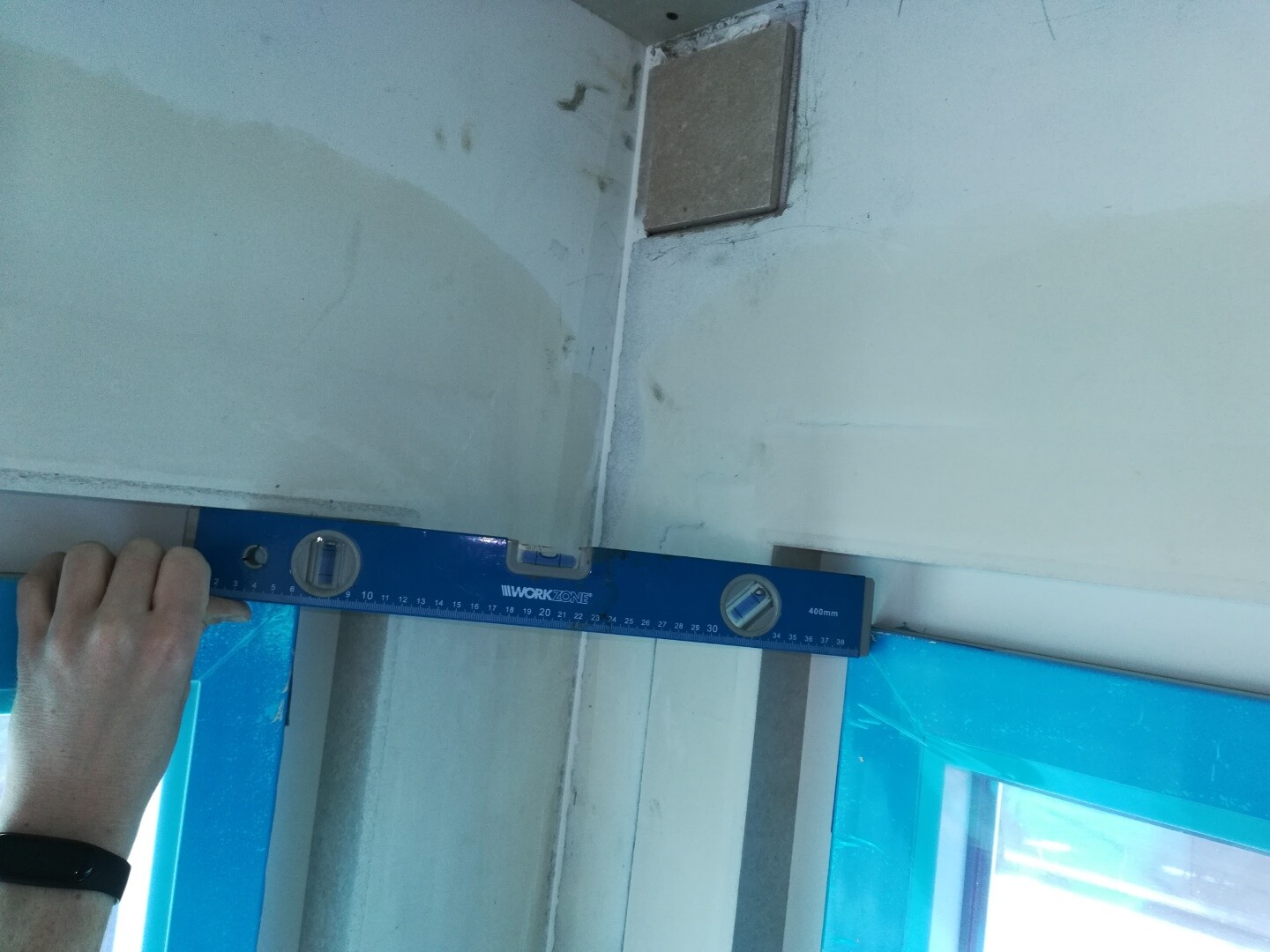 Eine Hand hält eine blaue Wasserwaage über Wandnaht in Bauraum, Fenster mit blauer Folie.