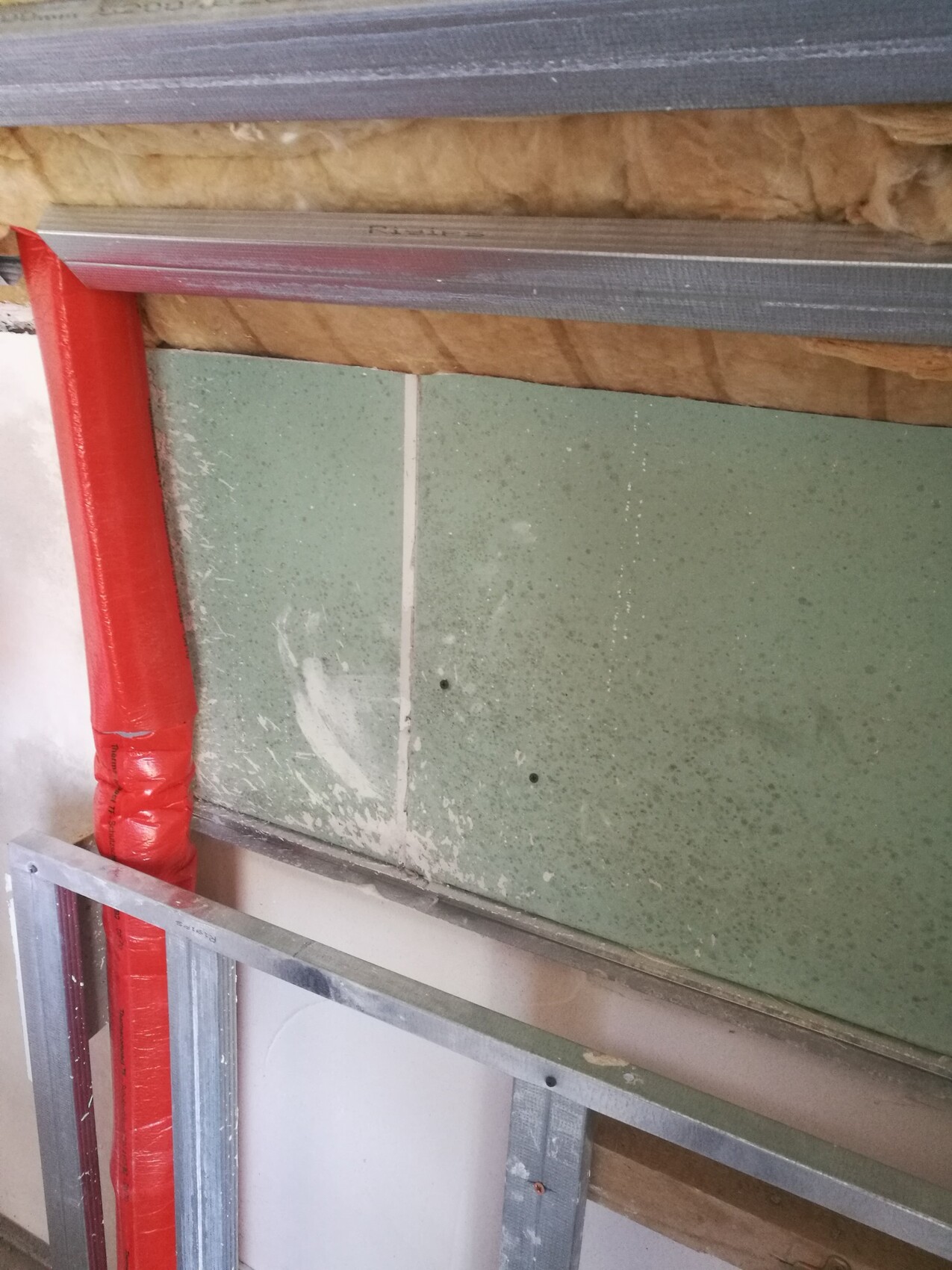 Sichtbare Rohbauwand mit Metallständern, Dämmung und roter Rohrleitung auf Baustelle.