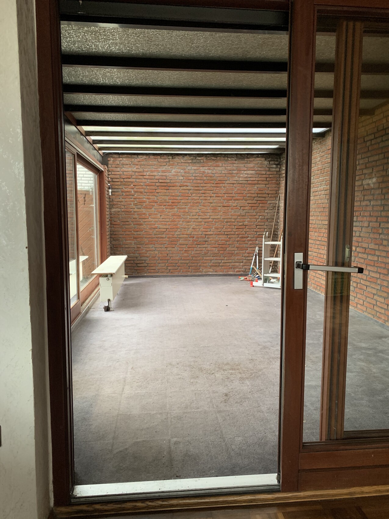 Verglaste Veranda mit Backsteinwänden, Holzrahmen-Tür; linke Fensterfront, Leiter im Hintergrund.