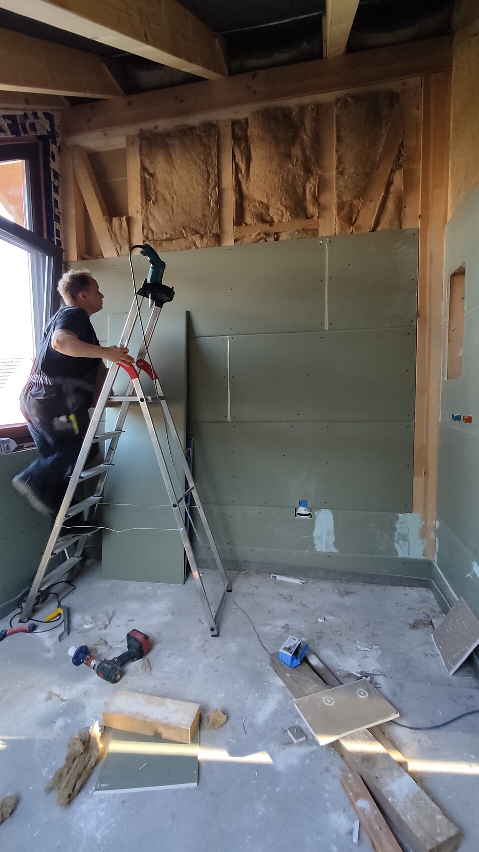 Mann auf Leiter montiert grüne Gipskartonplatten an einer Wand auf einer Baustelle.