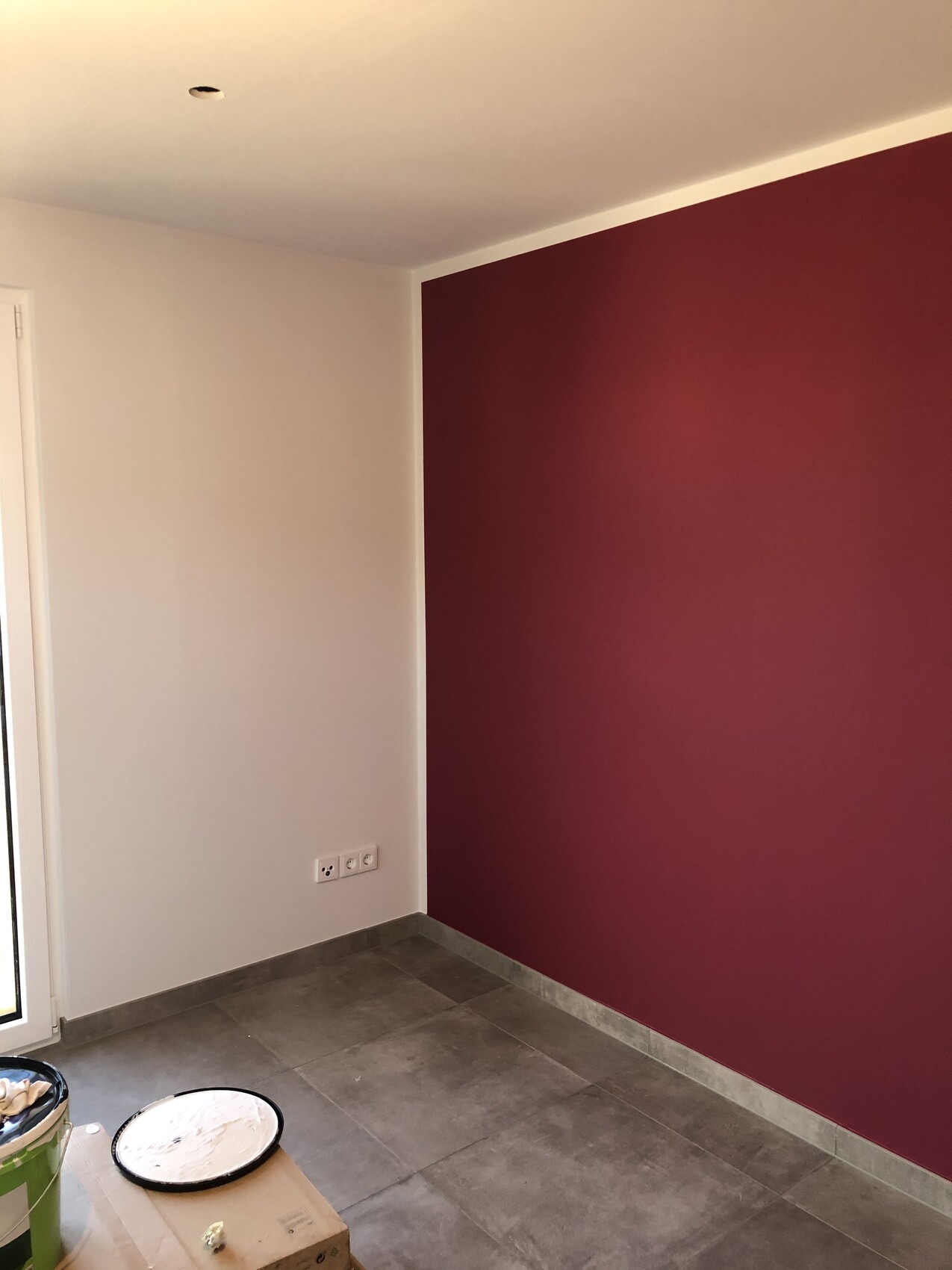 Zimmer-Ecke mit roter Akzentwand rechts, weiße Seitenwand, Fliesenboden, Renovierwerkzeuge.