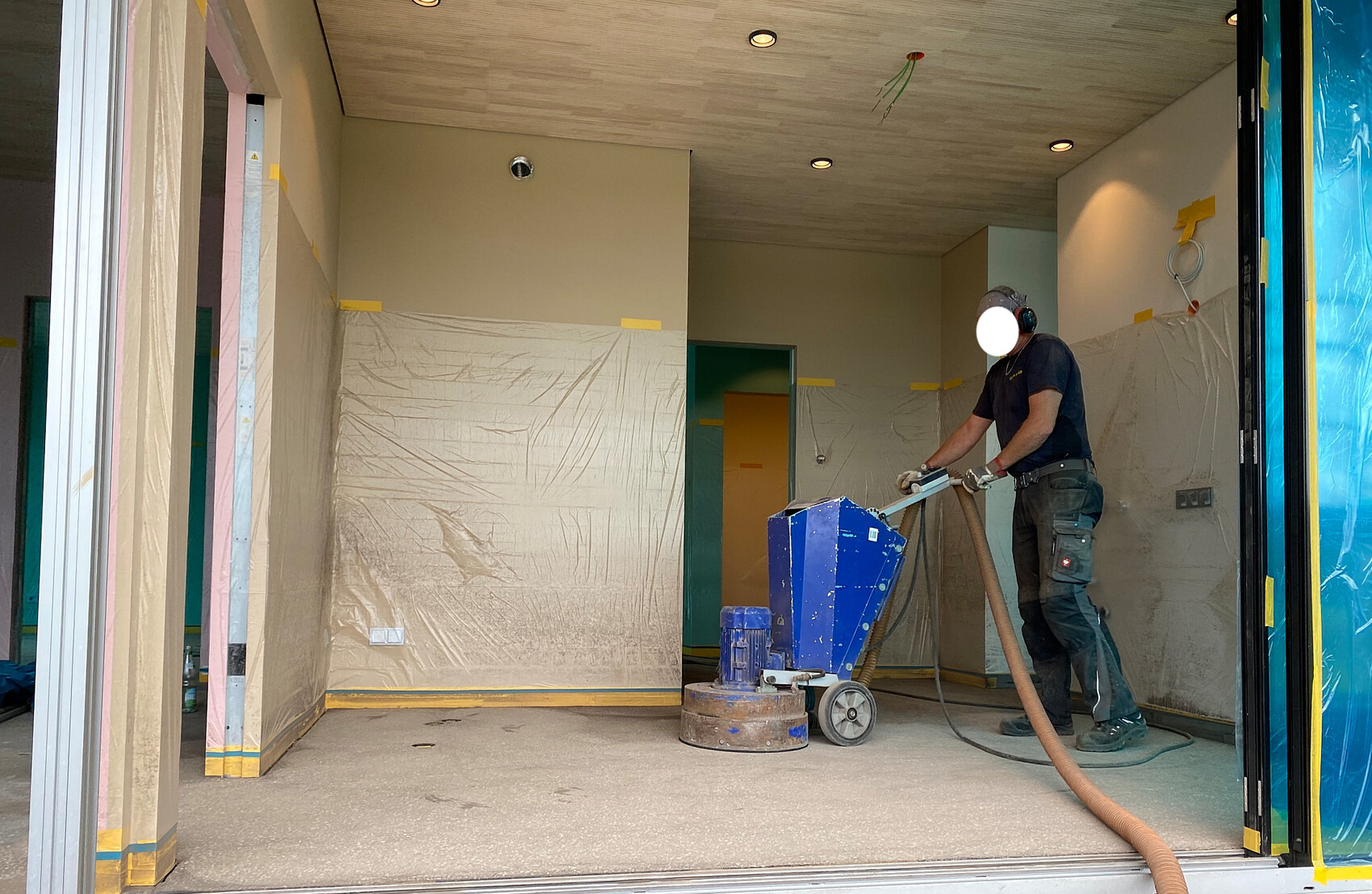 Bauarbeiter mit Gehörschutz schleift den Boden eines Innenraums, der mit Plastikfolien abgedeckt ist.