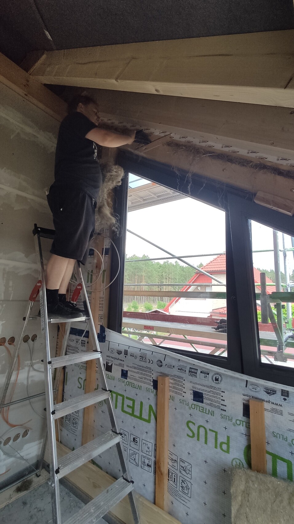 Person steht auf einer Trittleiter und arbeitet am Fensterrahmen in einer Baustelle im Dachgeschoss.