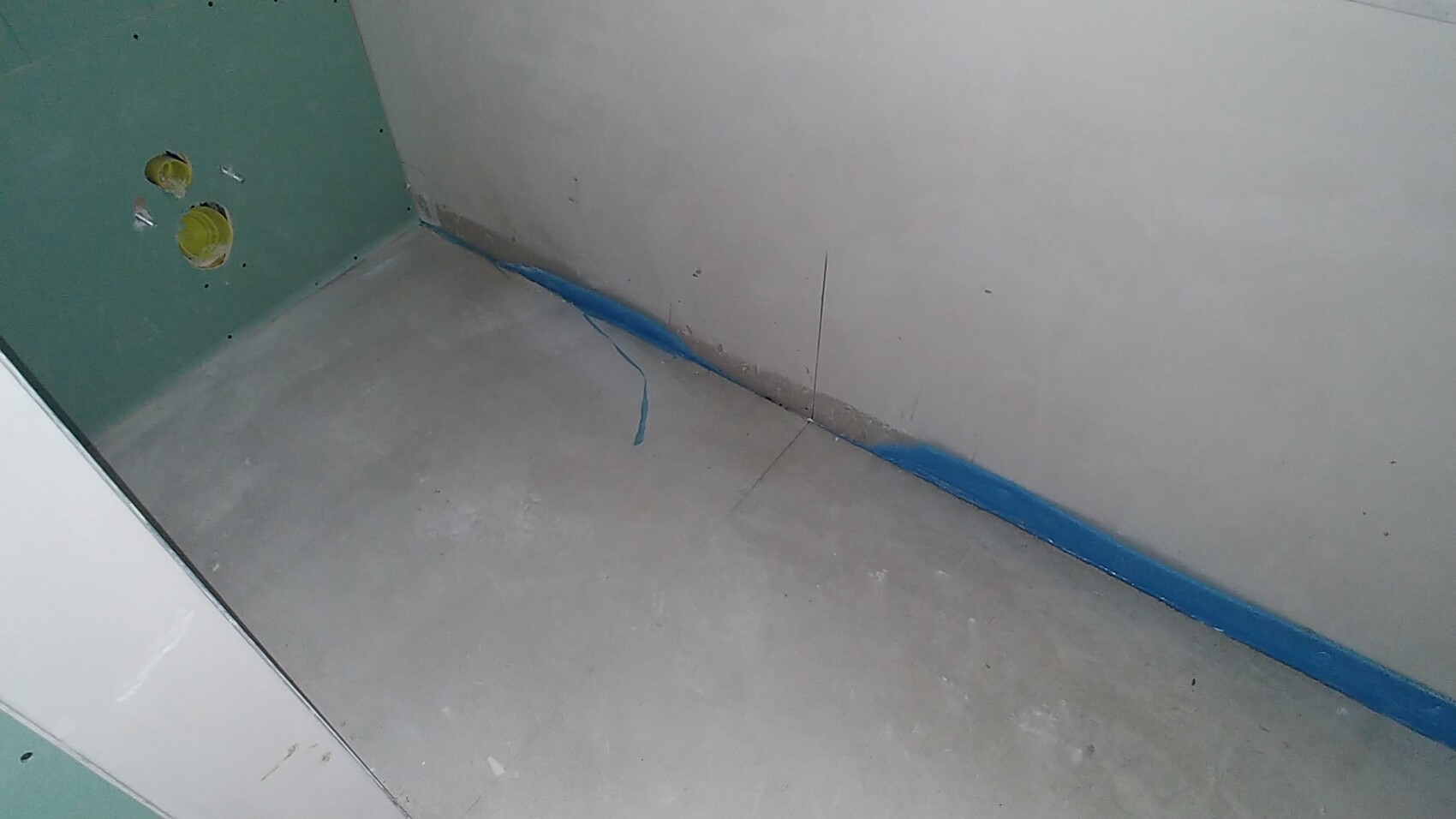 Unbearbeiteter Innenbereich im Bau: grüne Trockenbauwand, grob verlegter Estrich, blaues Dichtband am Boden.