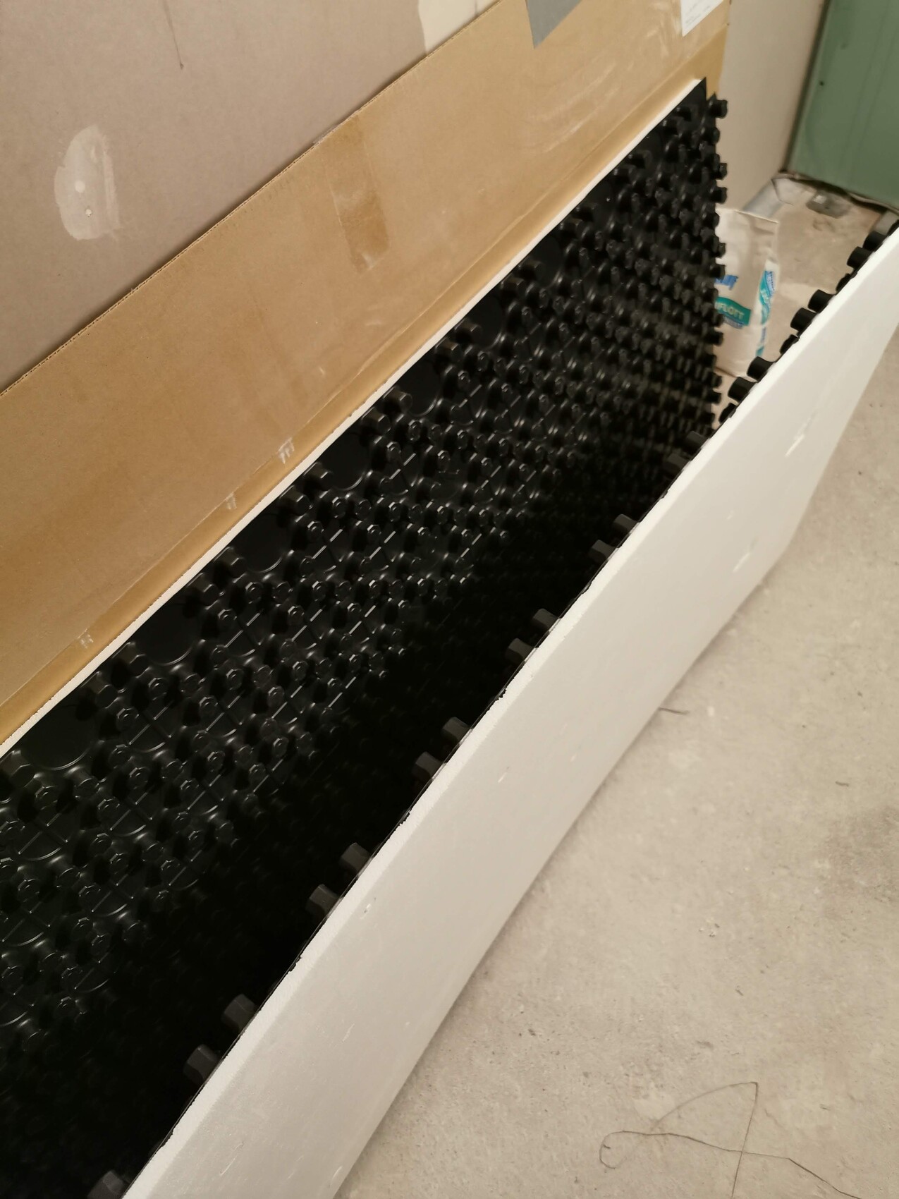 Weißer langer Behälter mit vielen schwarzen runden Stützen, Karton darüber, Betonboden.