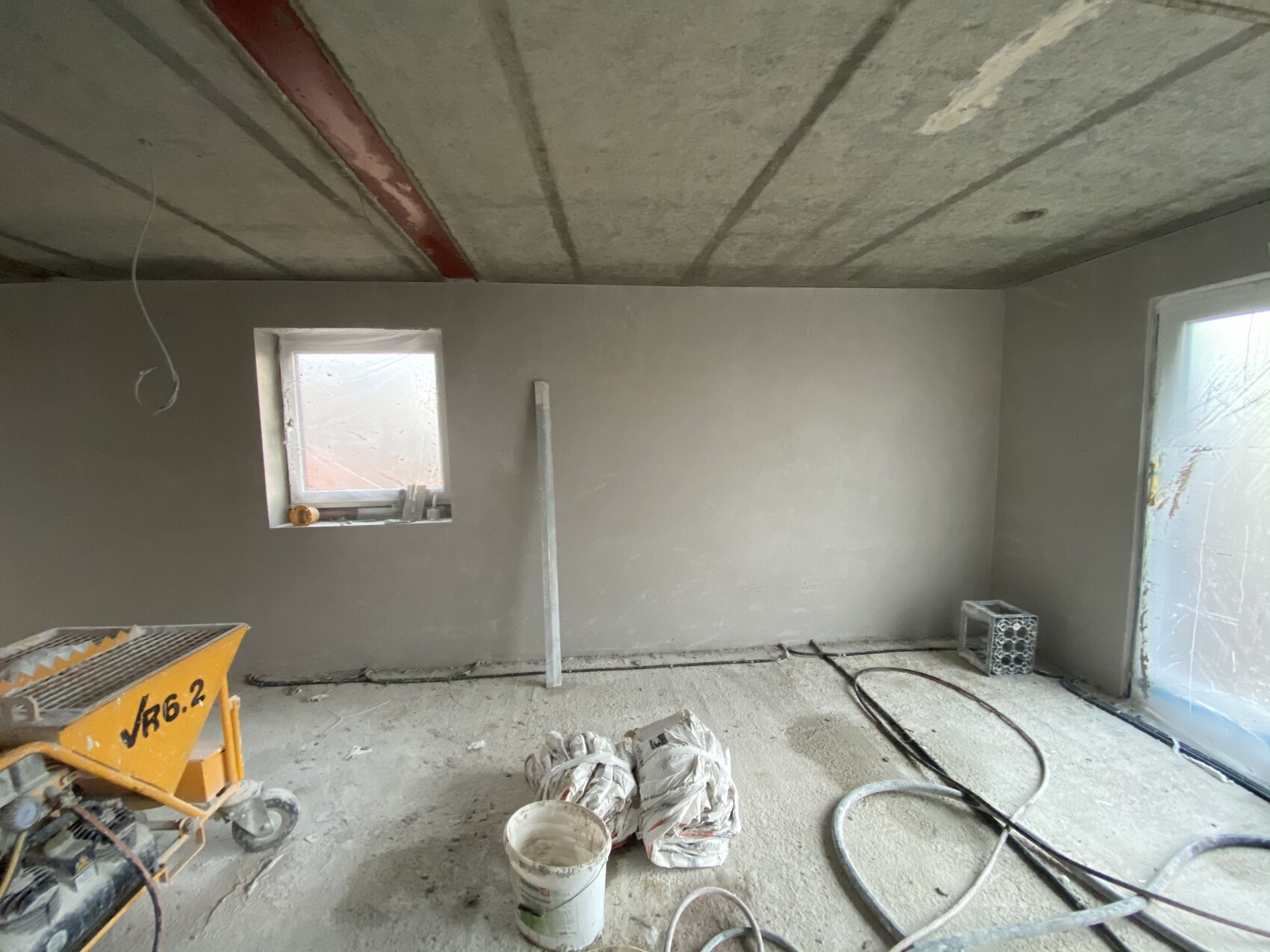 Rohbauzimmer: unfertige Wände/Decke, Bauwerkzeuge, Kabel, Eimer, Fenster links.