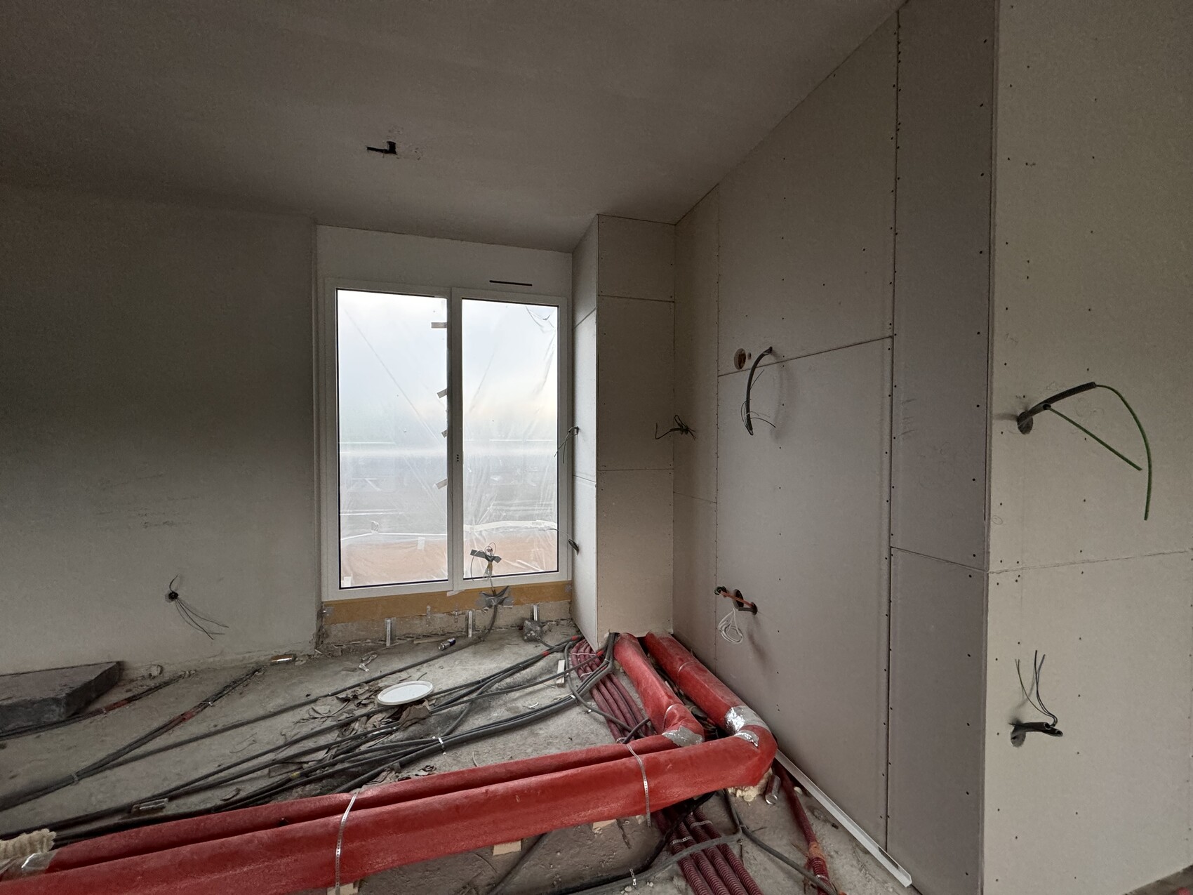 Rohbaustelle in einem Zimmer: unfertige Wände, lose Kabel, rote Rohre, Fenster mit Ausblick.