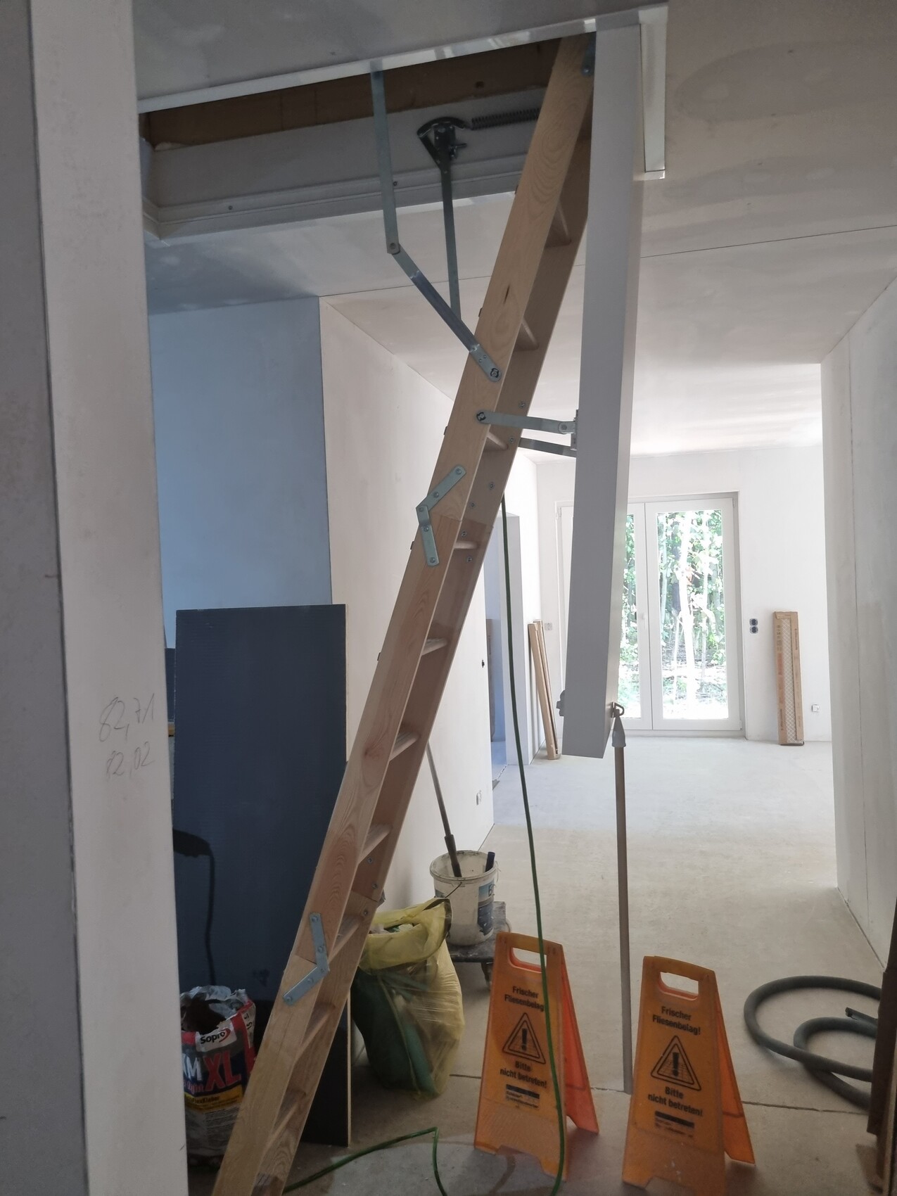 Baustellenraum mit Holzleiter gegen Decke, Eimer, Säcke und Warnschilder; Fenster am Ende.