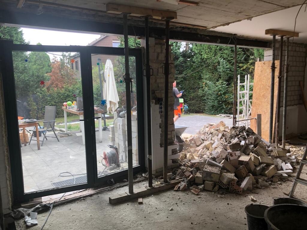 Bau-Raum mit Ziegelresten und Stützen; Glas-Schiebetür zum Garten; draußen Tisch und Stühle.