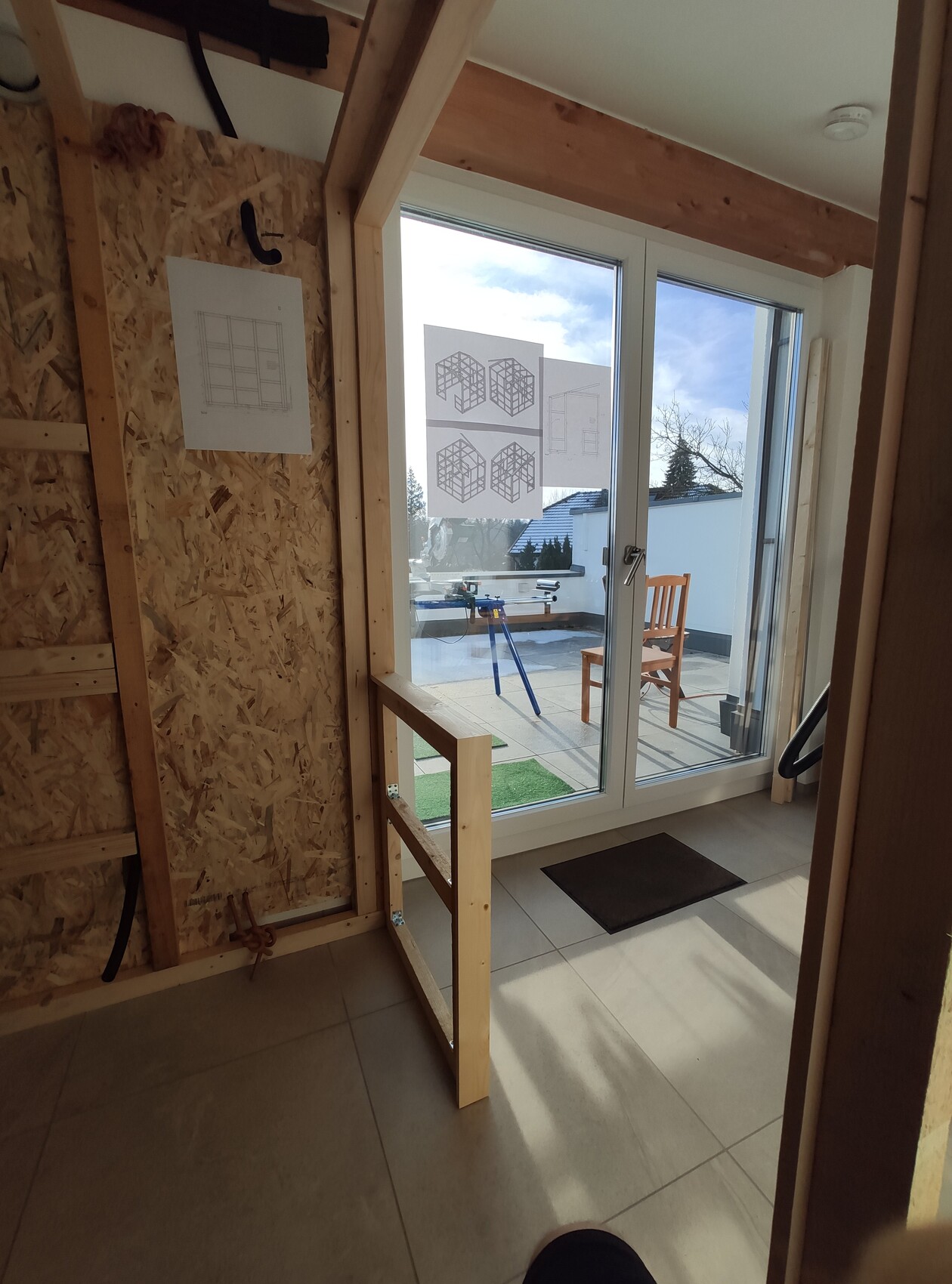 Innenansicht einer Baustelle: Holzrahmen, OSB-Wand, Glas-Tür zum Außenbereich, Baupläne an der Wand.