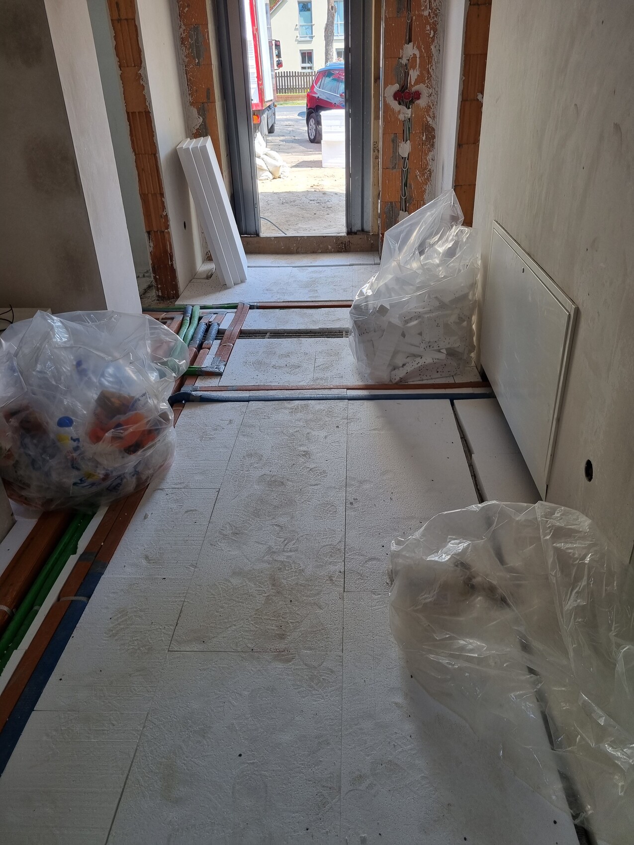 Flur einer Baustelle mit Plastiksäcke, Rohren, Platten und offener Tür nach draußen.