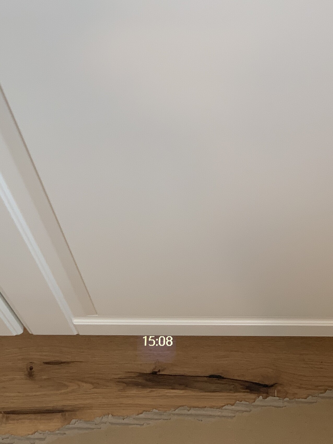 Weiße Wand mit Türrahmen links, Holzboden, beschädigte Bodenkante; Uhr 15:08 projiziert