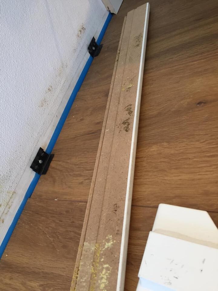Lange Holzleiste auf Laminatboden neben weißer Wand; blaues Malerband und schwarze Halterungen.