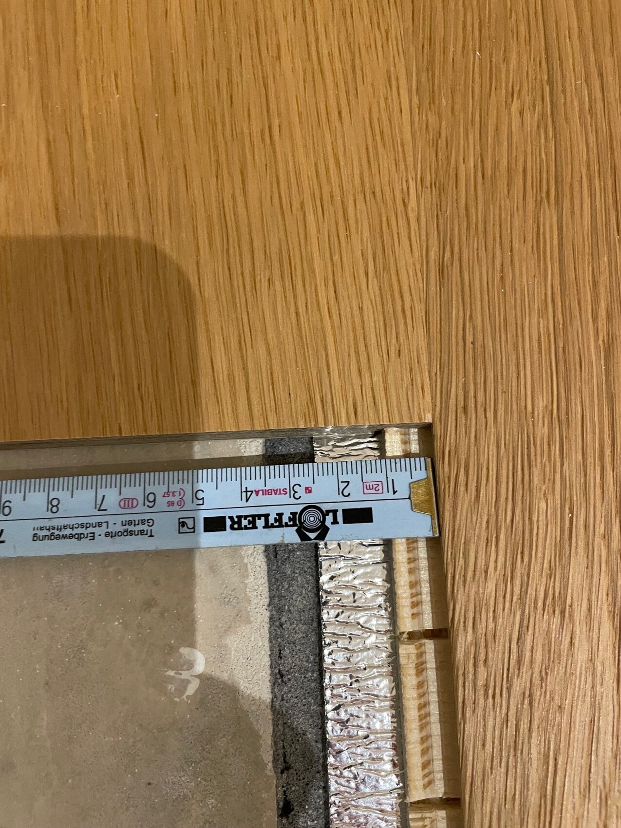 Maßband liegt quer über eine Holzoberfläche neben einem Rand aus Aluminiumfolie und Dämmmaterial.
