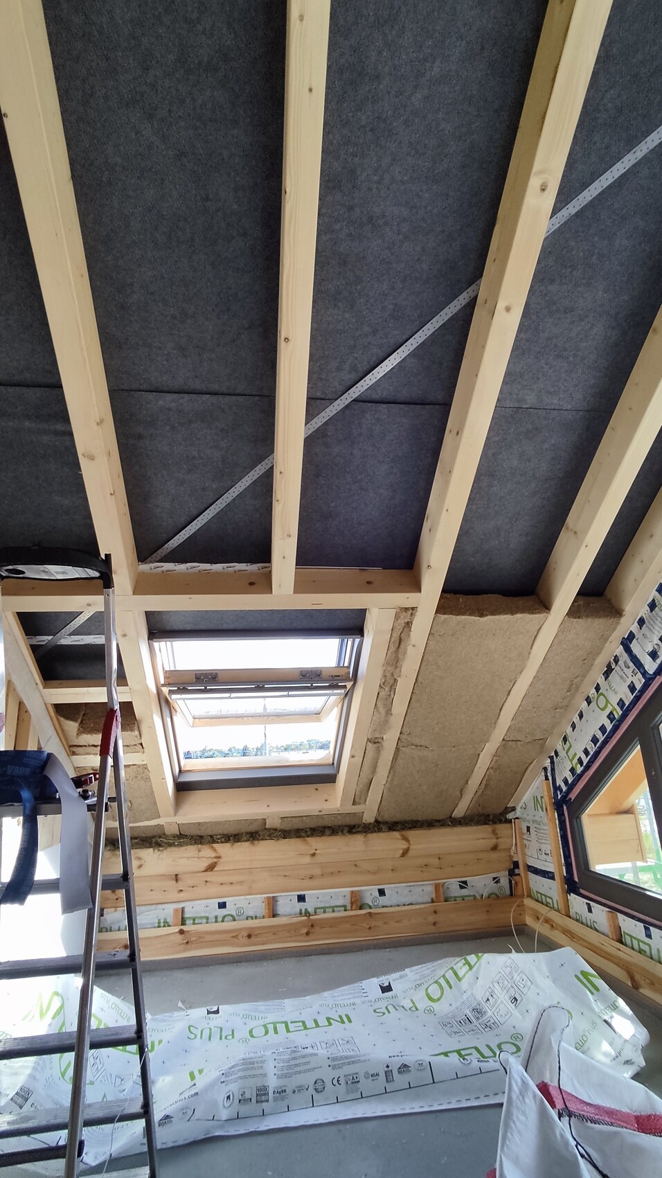 Baustellenraum mit Holzbalken-Decke, Dachfenster, Leiter, Dämmung und Baufolie.
