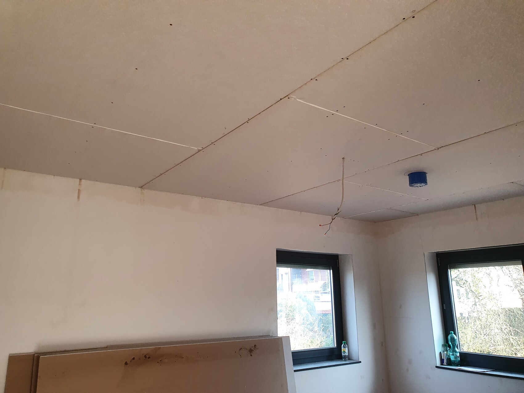 Unfertiger Raum: Deckenplatten, sichtbare Fugen, lose Kabel, blauer Anschlussdeckel, zwei Fenster.