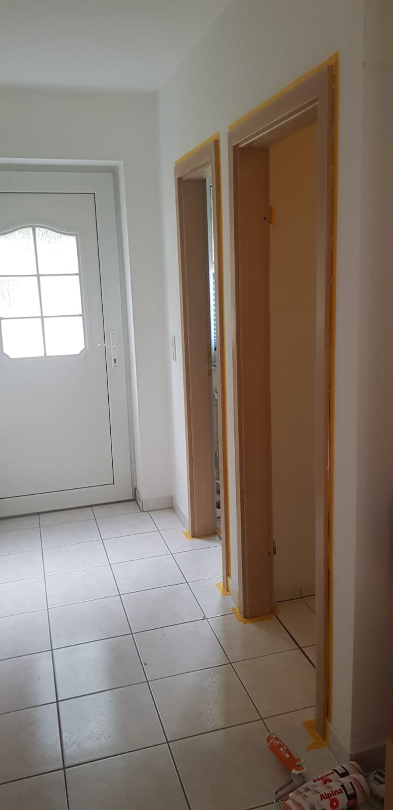 Flur mit weißer Haustür, daneben zwei unfertige Türrahmen mit gelbem Malerband; Fliesenboden.