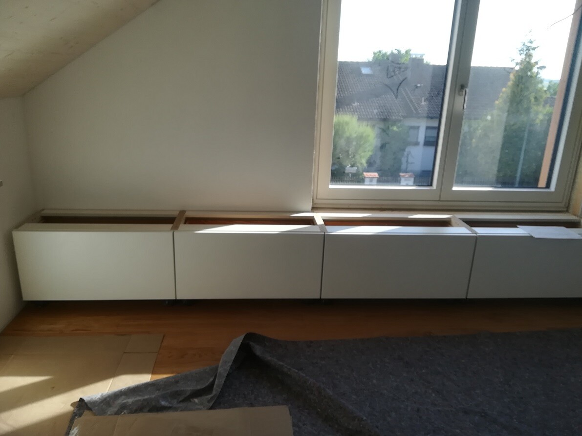 Heller Raum mit vier weißen Unterschränken unter großem Fenster, Holzboden.