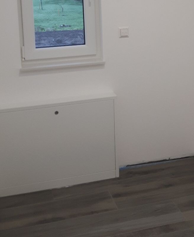 Weiße Innenwand mit kleinem Fenster über einer weißen Schrankbank, dunkler Boden, blaues Malerband.