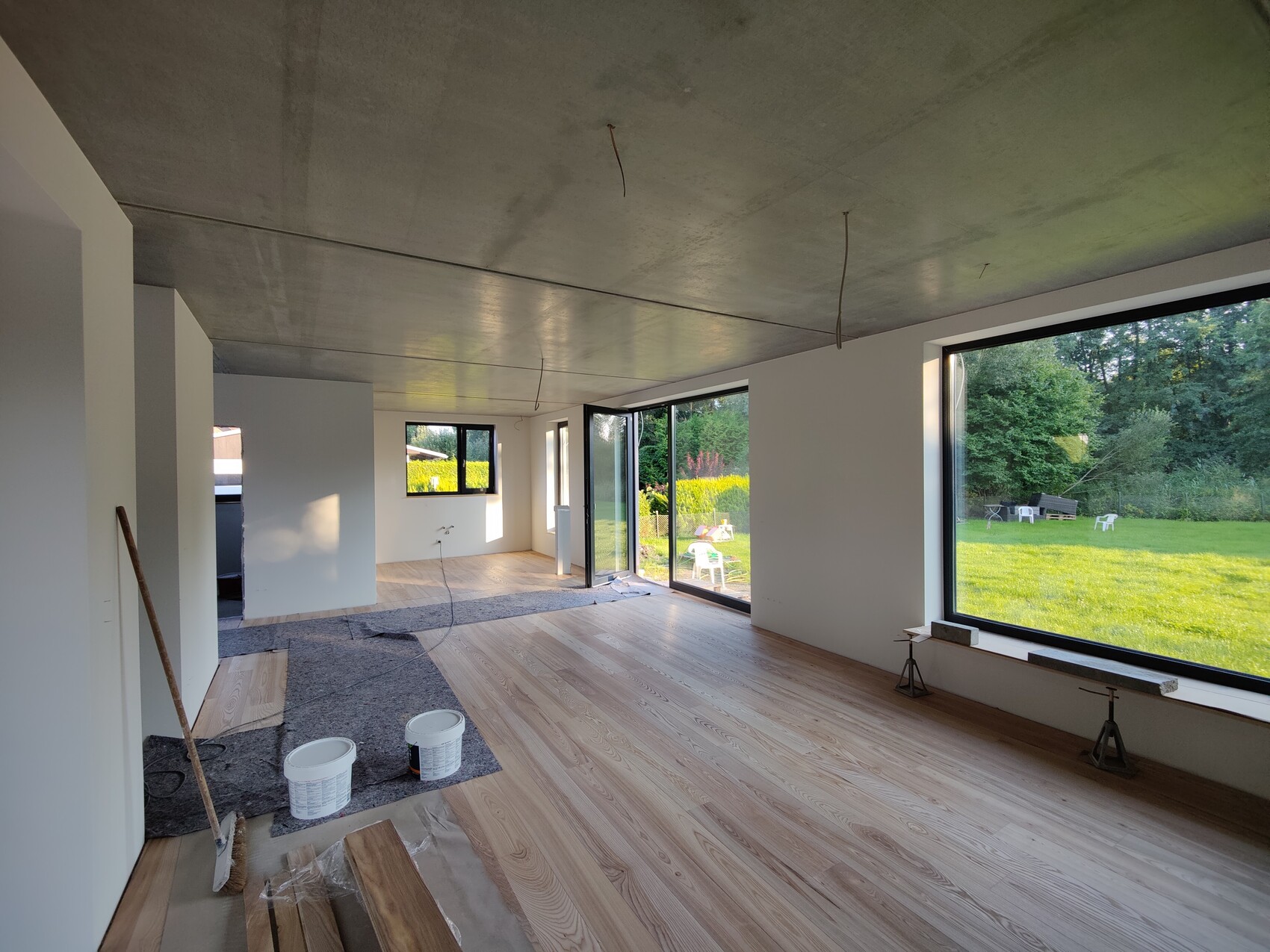 Großer Bauinnenraum mit roher Decke, Holzfußboden, Eimern, Besen; Blick auf Garten durch Fenster.