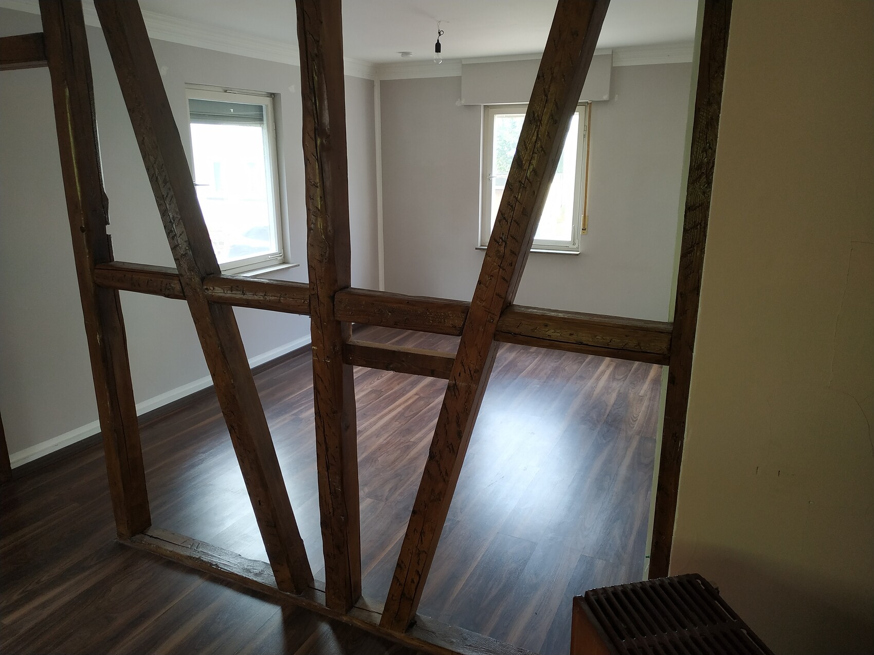 Innenraum mit freigelegten Holzbalken und Ständern, dunkler Holzfußboden, zwei Fenster.