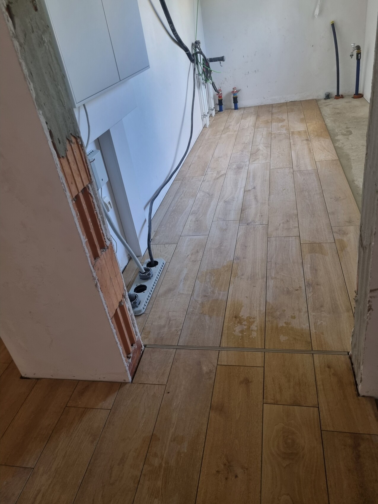 Waschraum links mit Waschmaschine an der Wand, Holzbodenplatten, Schläuche und Wasserhähne.