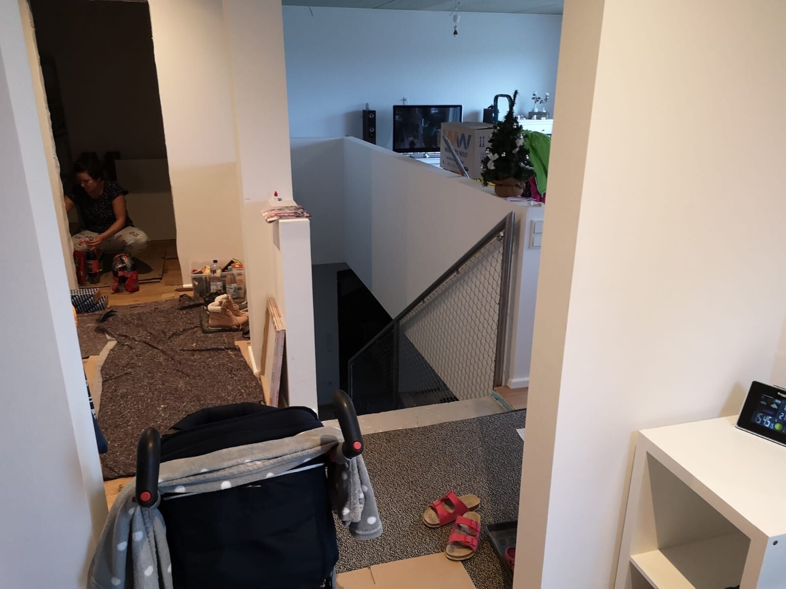 Wohnbereich im Umbau: linke Person sitzt mit Bastelmaterial, Kinderwagen vorne, Treppe rechts.