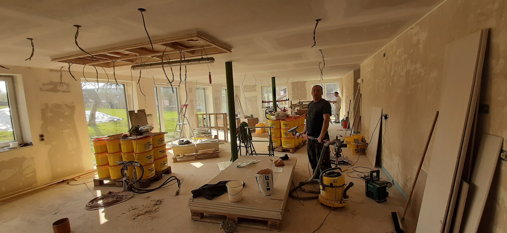 Innenraum einer Baustelle: Mann mit Werkzeug, gelbe Eimer, hängende Kabel von der Decke.