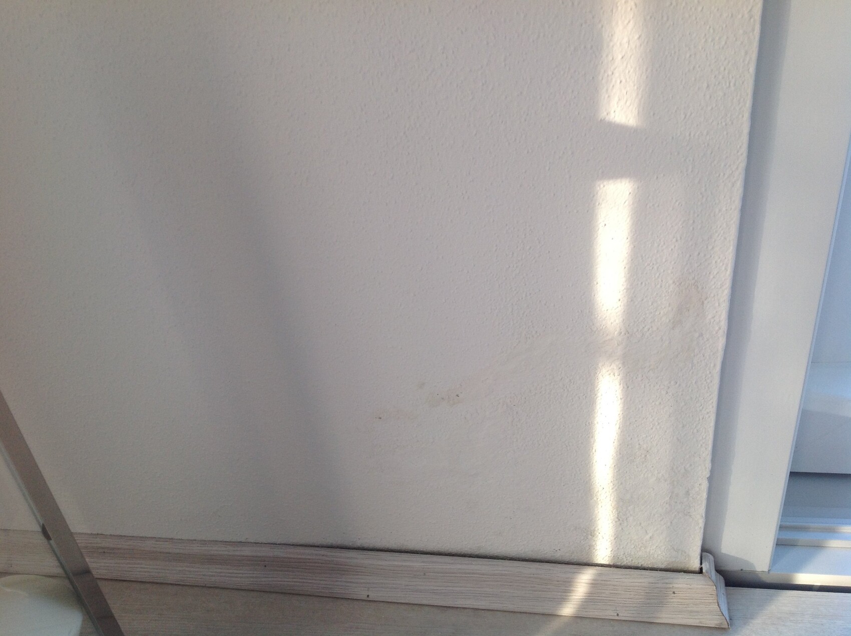 Weiße Wand mit Textur im Innenraum, Holzsockelleiste unten, rechter Türrahmen, Lichtreflexe.