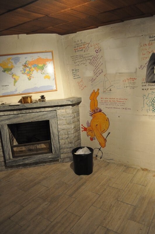 Innenraum mit Kamin, Weltkarte an der Wand und orange Comicfigur; Mülleimer im Vordergrund.