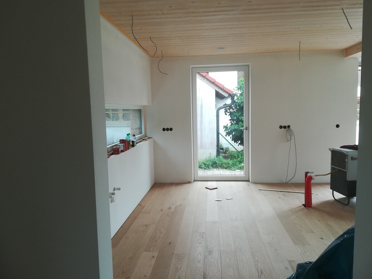 Küche im Bau: Weiße Schränke, Holzboden, offenes Kabelgewirr, Tür zum Garten sichtbar.