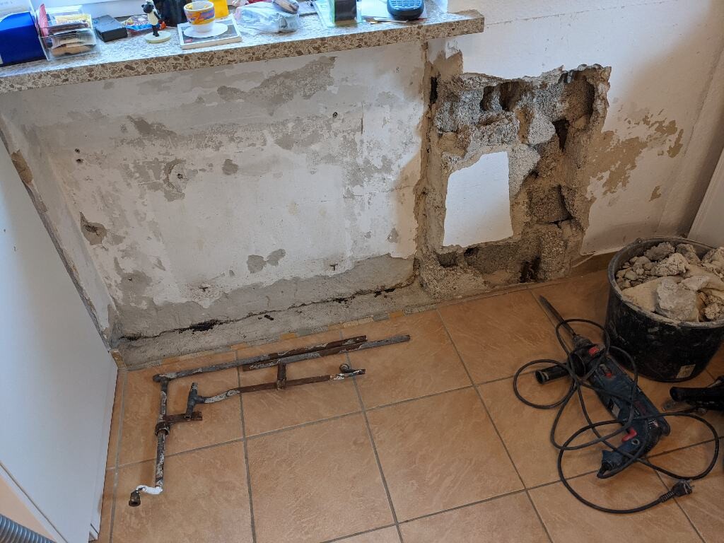 Küchenwand aufgerissen, freiliegende Rohre, Schutt, Werkzeuge und Eimer am Boden.