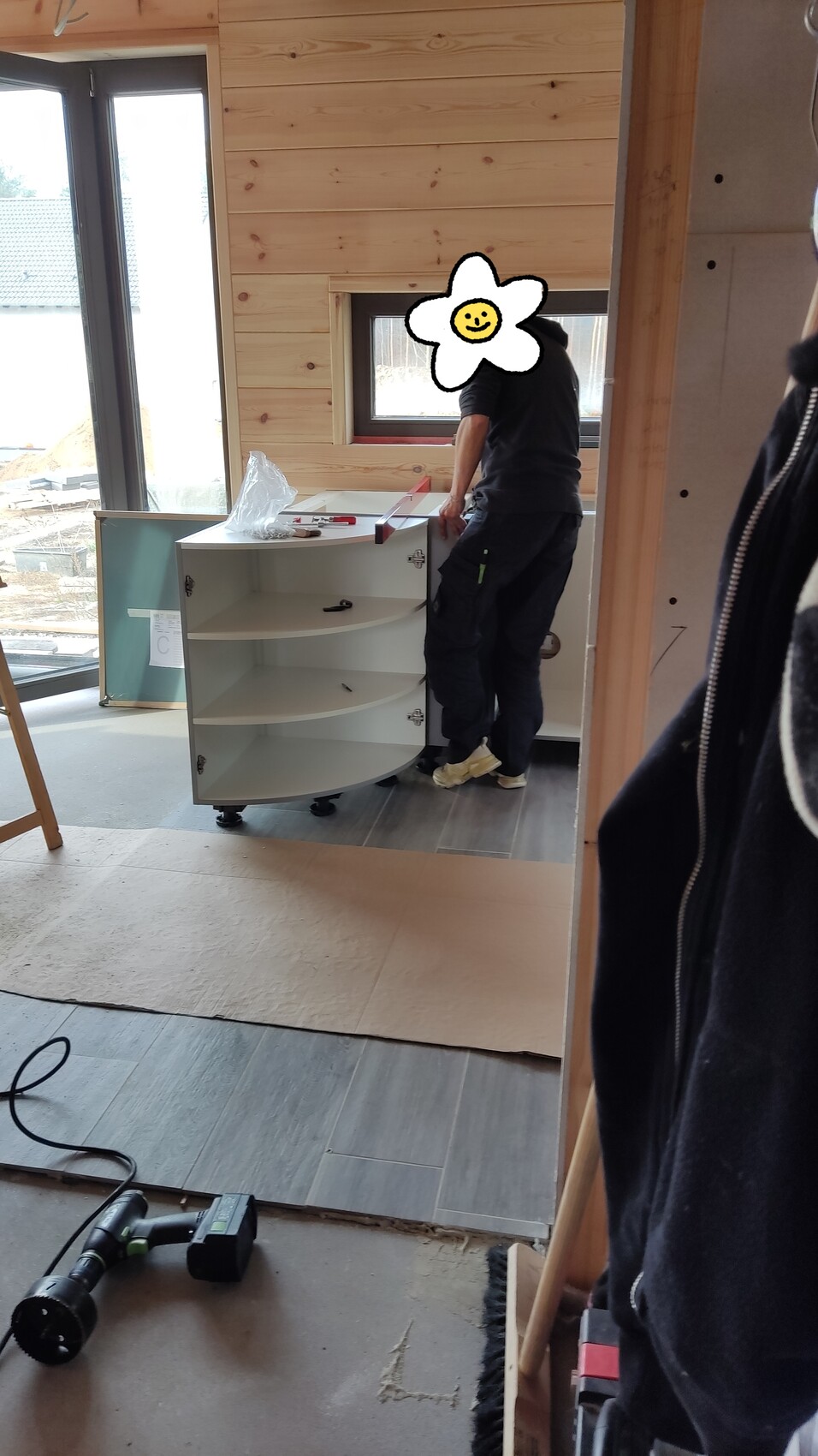 Person montiert einen weißen Eckschrank in einem Holzbau-Raum; Werkzeuge liegen auf dem Boden.