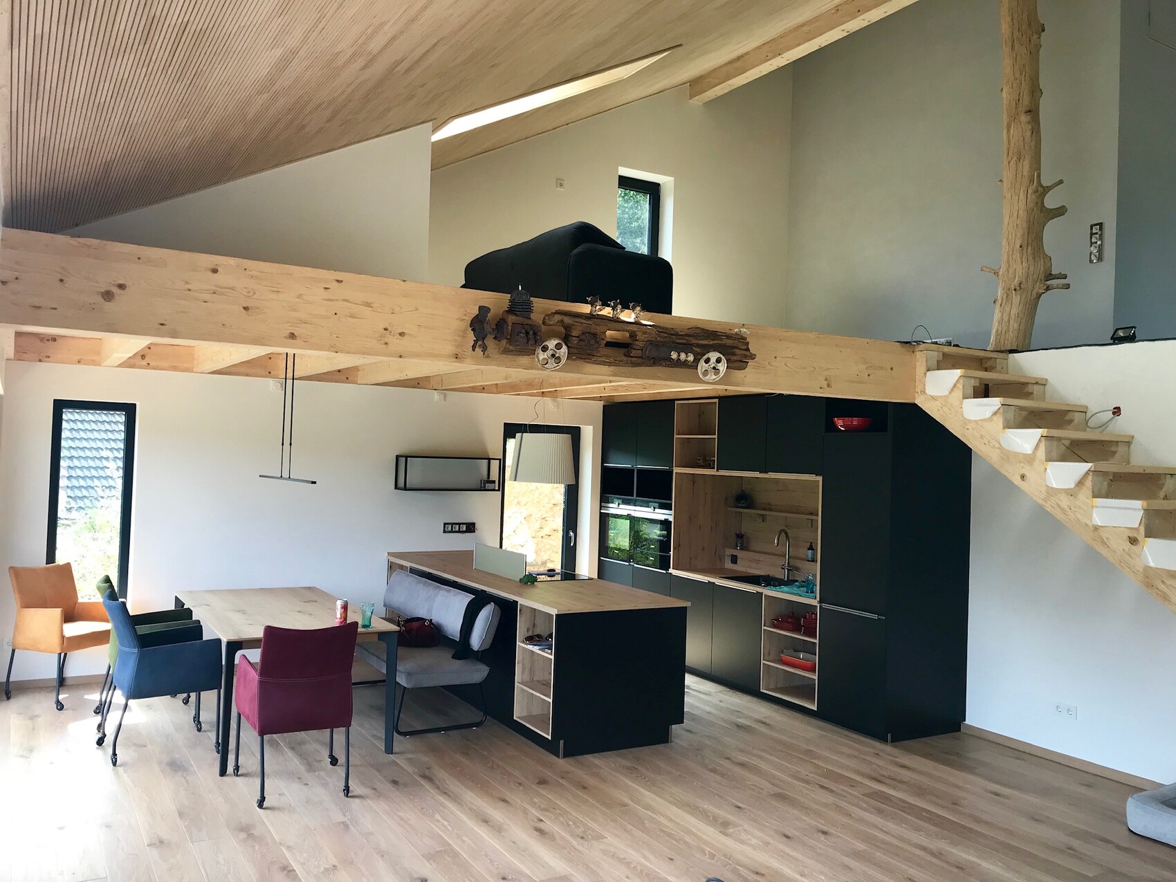 Offener Innenraum mit Küche, Esstisch, bunten Stühlen und Loft-Galerie aus Holz.