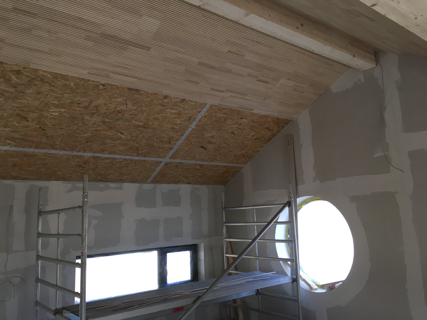 Baustellenraum mit OSB-Decke, Gerüst, grauen Rigipswänden und rundem Fensterloch.