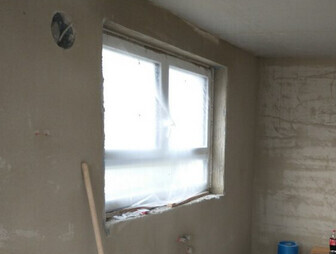 Unbearbeiteter Innenraum während Renovierung; Fenster abgedeckt, rohe Betonwände sichtbar.