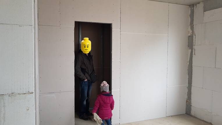 Erwachsener in dunkler Jacke mit LEGO-Kopf steht in Türöffnung; Kind in pinker Jacke nähert sich
