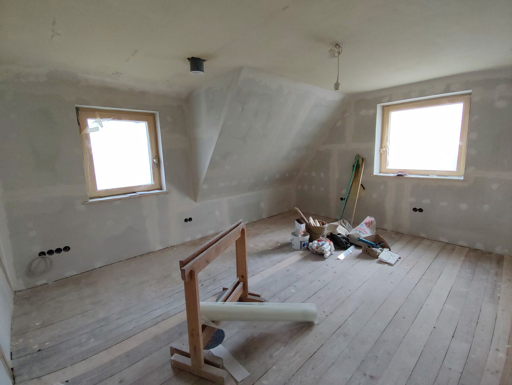 Renovierungsraum mit unfertigen Wänden, zwei Fenster, Werkzeugen und Baumaterialien auf Holzboden.
