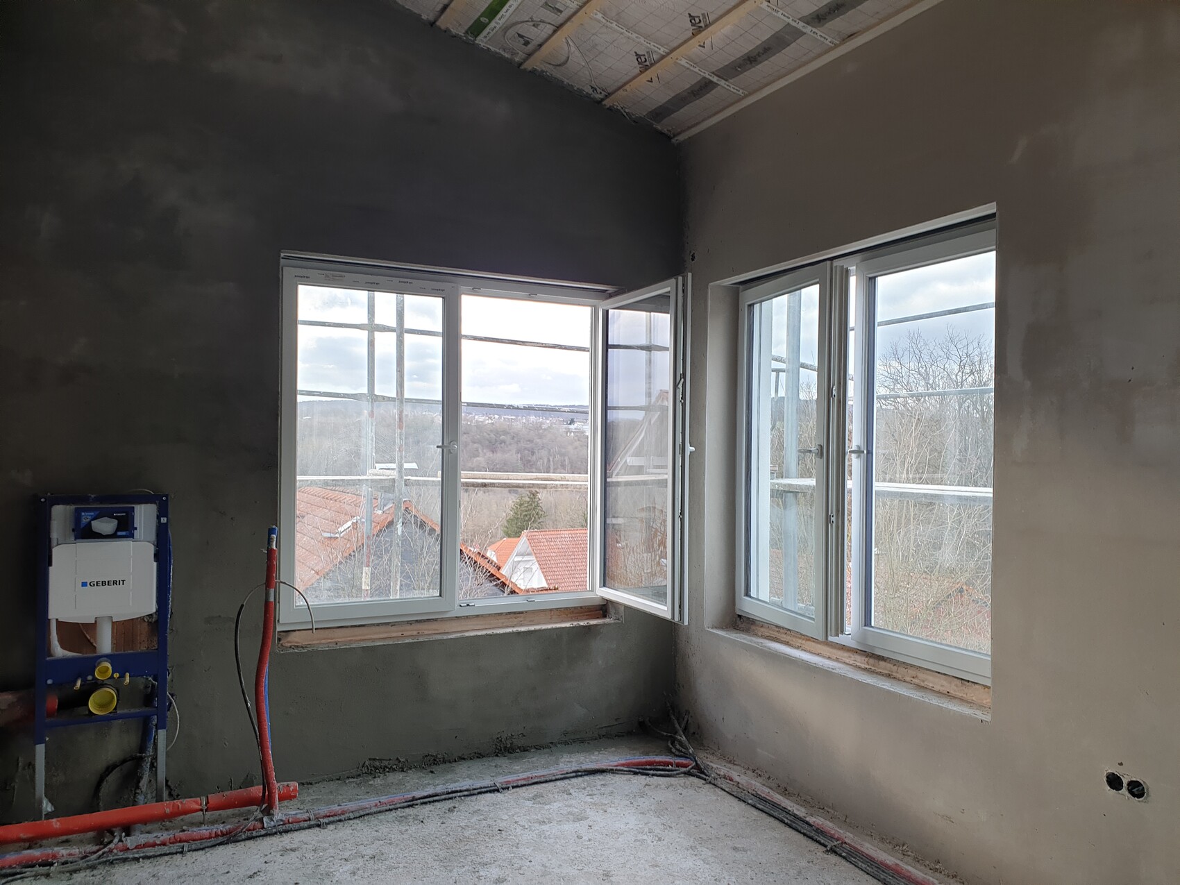 Unfertiges Zimmer mit zwei großen Fenstern, sichtbare Rohre, Bauinstallation, Aussicht nach außen.