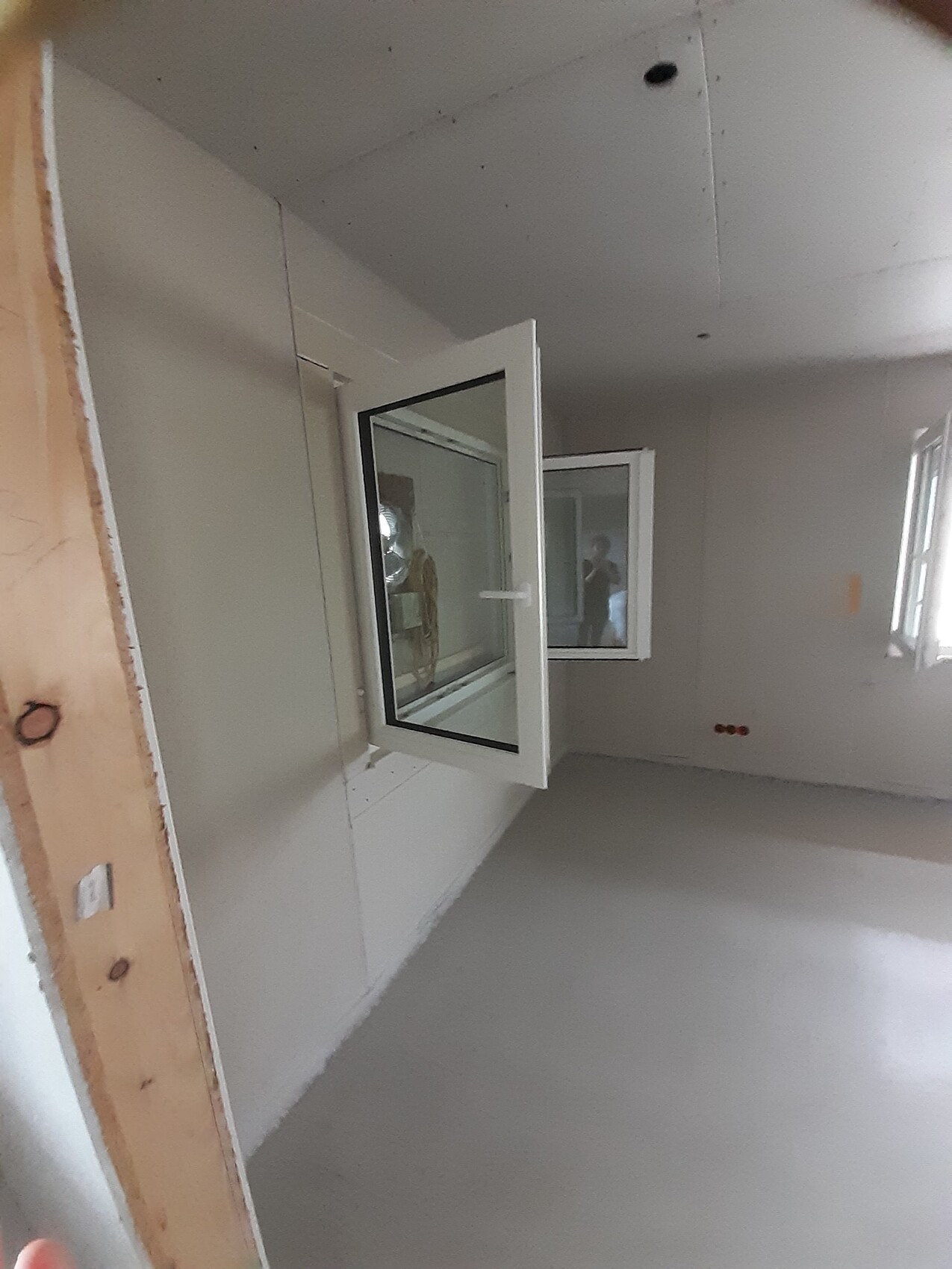 Unfertiger Innenraum mit geöffnetem weißen Fenster, glatten Wänden und Bauholz links.