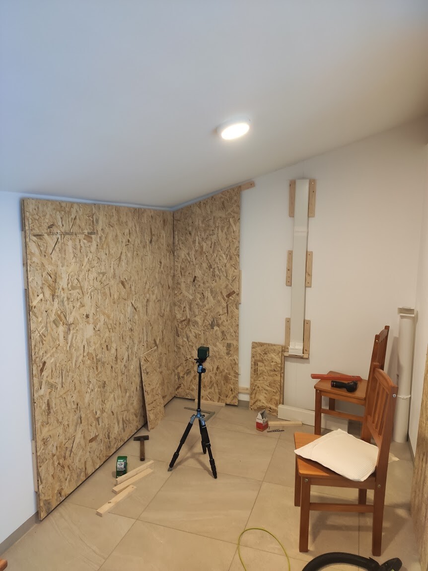 Innenraum-Werkstatt mit OSB-Wänden, Stativkamera, Holzplatten und zwei Stühlen.