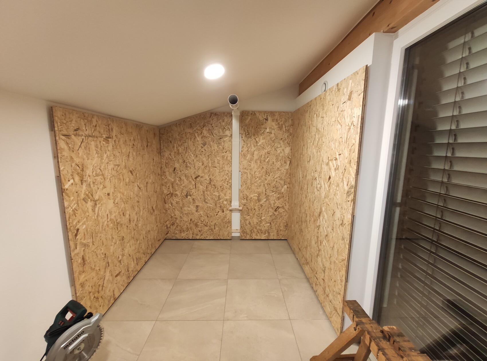 Kleiner Innenraum mit OSB-Wänden, Deckenleuchte, Rohr, Fenster mit Jalousien und Bauwerkzeugen