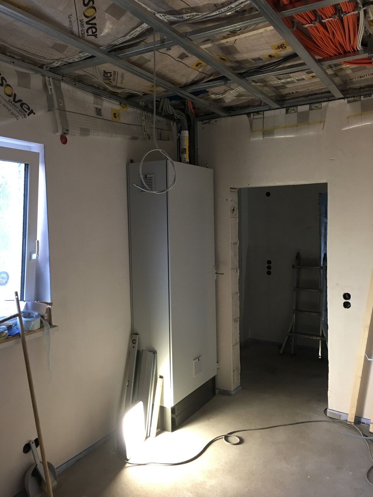 Unfertiger Innenraum mit Installationsschrank, offener Decke, Kabelwirrwarr, Leiter und Fenster.